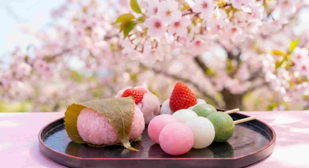 A plate of sakura sweets including pink sakura mochi and sanshoku hanami dango under blooming cherry blossom trees.