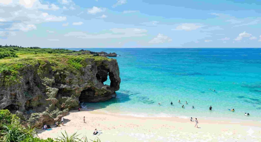 A large natural limestone rock archway covered in green vegetation stands at the edge of a pristine white sand beach. The ocean water fades from clear shallow water to deep turquoise, with people swimming and relaxing on the shore.