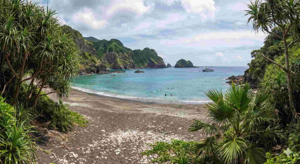 A secluded beach with dark volcanic sand and scattered white coral, framed by lush green tropical vegetation. Turquoise water laps the shore where people are snorkeling, with rugged, green cliffs and a boat in the distance under a partly cloudy sky.