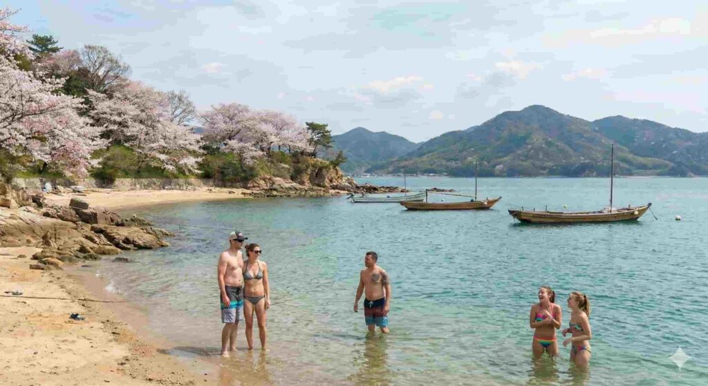 A small group of Western tourists standing and relaxing in calm, clear turquoise ocean water. On the left shore, pink cherry blossom trees are in full bloom above a rocky coastline. Traditional wooden fishing boats are anchored in the distance against a backdrop of green mountains and blue sky.