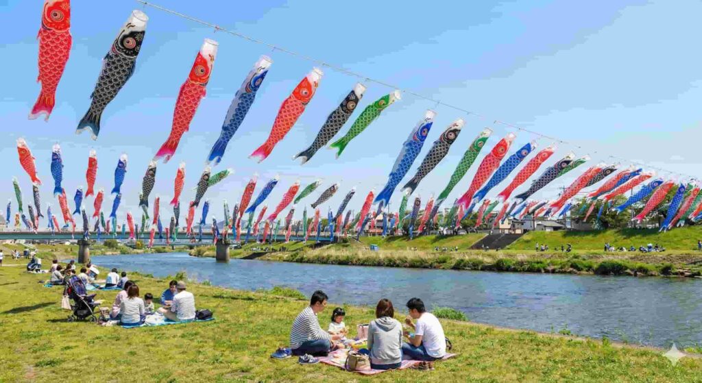 Hundreds of colorful Koinobori carp streamers flying across a blue sky over a riverbank with people picnicking below.