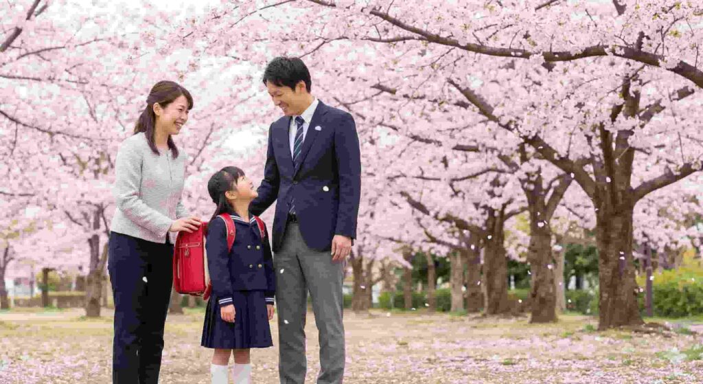 Japanese family with a young daughter in a school uniform and red backpack standing under blooming cherry blossom trees during spring.