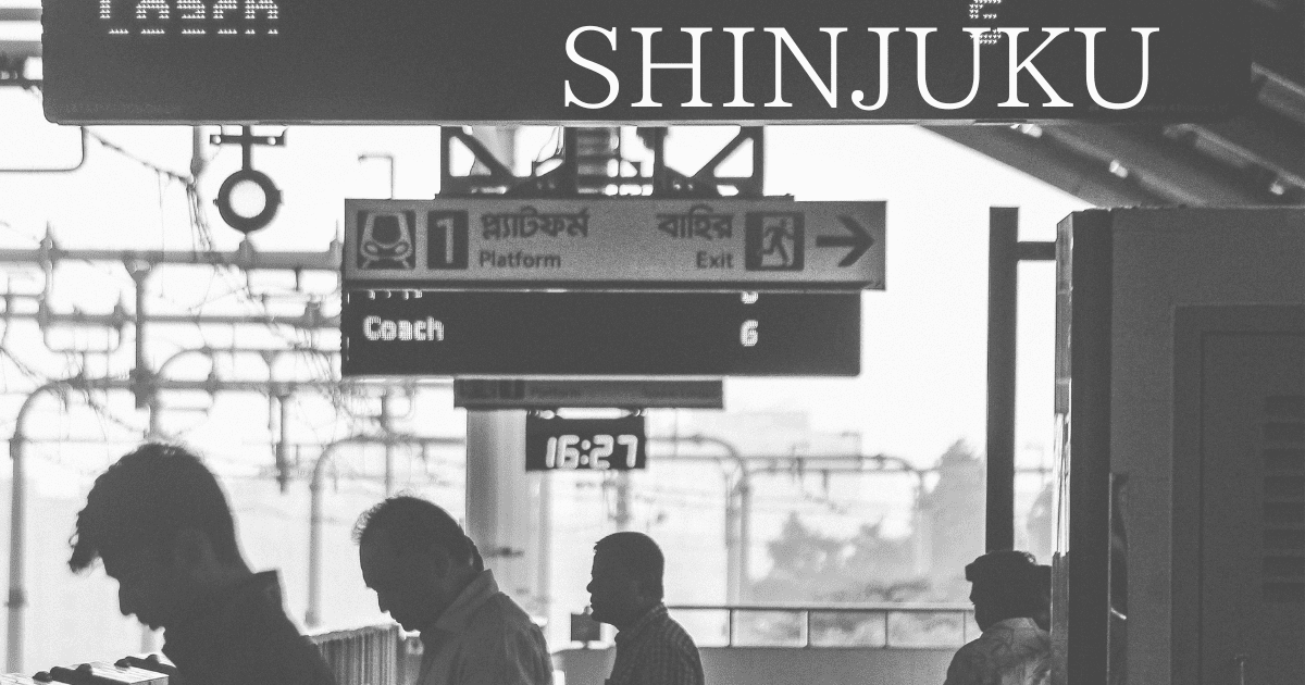 A black and white photograph captures a train platform with overhead signs. The largest sign reads "SHINJUKU" in white letters. Below it, another sign indicates "Platform 1" and "Exit" with an arrow, including text in Bengali. A digital clock displays "16:27". Several people in silhouette wait on the platform under a metal canopy and overhead wires.