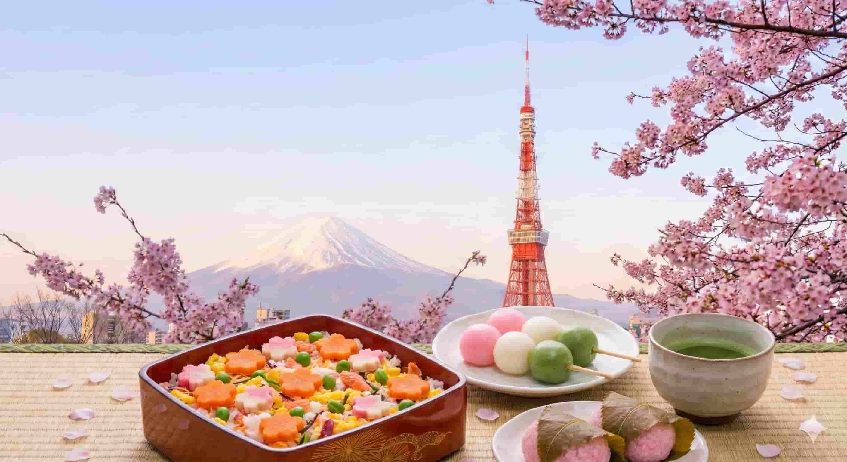 A traditional Japanese hanami picnic spread featuring a colorful chirashizushi bento box, sanshoku dango, and sakura mochi, set against a backdrop of blooming cherry blossoms framing Tokyo Tower and Mount Fuji at sunrise.